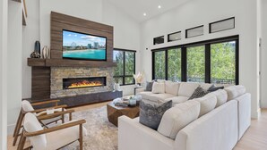 A modern living room with a stone fireplace, a large flat-screen TV, a white sofa, and large windows offering a view of greenery. - 6 Bedroom Ski In/Ski Out Luxury 6 Bedroom Private Gated Community (Park City)