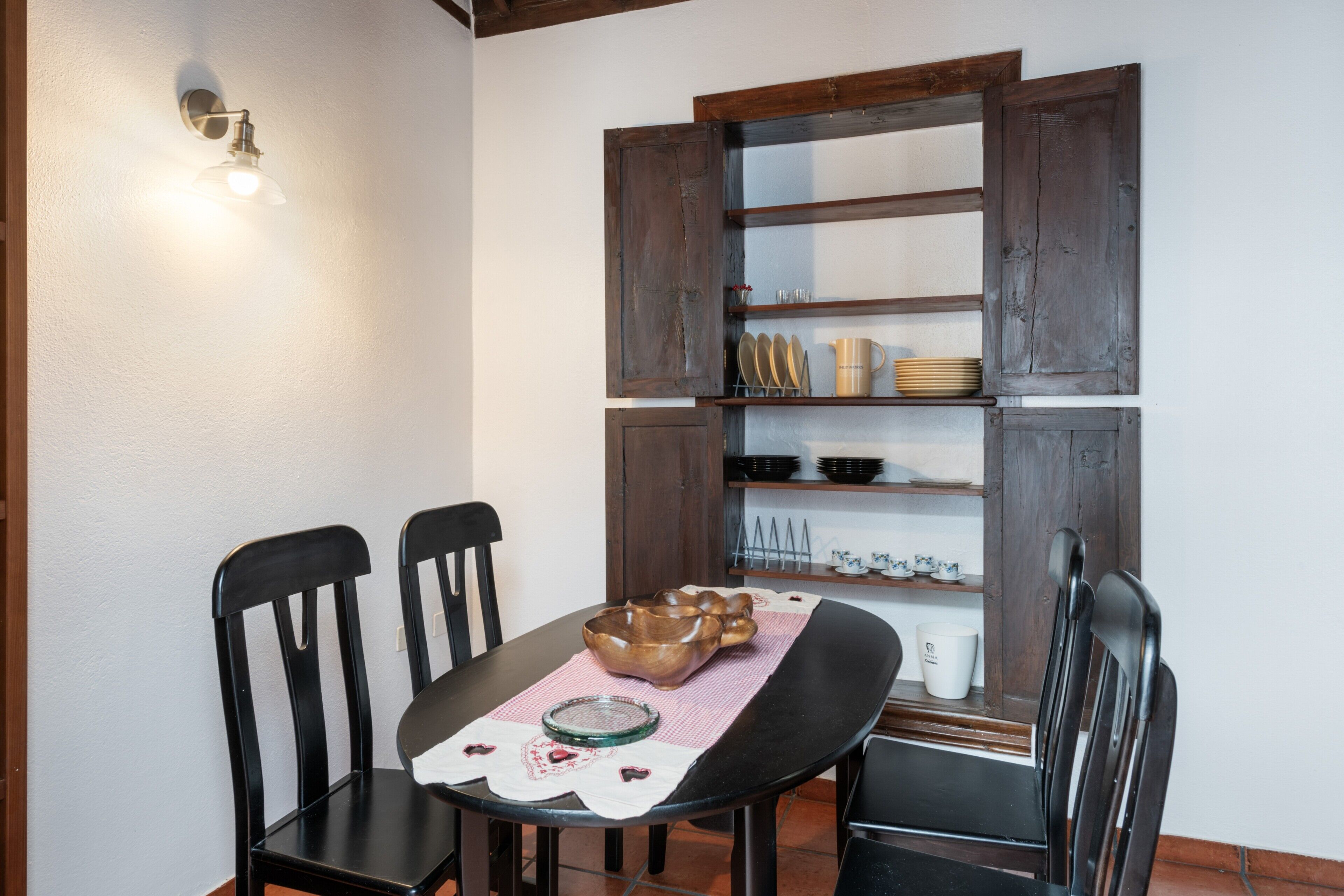 A dining table with chairs, a wooden cabinet with shelves, and a wall-mounted light fixture.