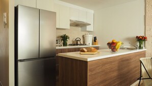 A modern kitchen with a stainless steel refrigerator, wooden cabinetry, a white countertop, and a small island with a cutting board and fruits.