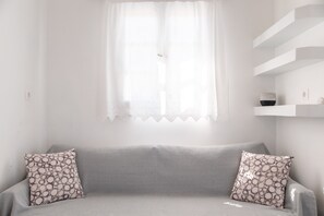 A neutral-colored sofa with patterned cushions, a white window with a sheer curtain, and a white shelf with a decorative item.