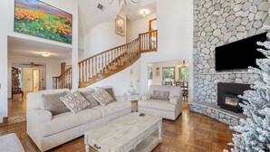 A spacious living room with a wooden staircase, a stone fireplace, and a large TV mounted on the wall. - Vineyard Vista Retreat– Your Serene Getaway in Temecula (Temecula)