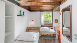 Ett litet rum med ett träpanelat tak, ett fönster med utsikt över grönområden, en säng med en kudde, ett litet skrivbord med en stol och en hylla med dekorativa föremål.