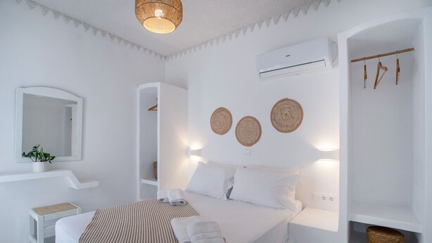 A white bedroom with a bed, bedside table, and a mirror. There is an air conditioning unit on the wall and a shelf with baskets.