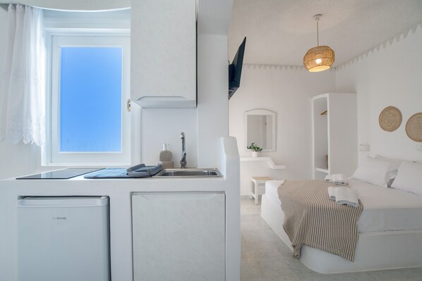Une chambre compacte et moderne, dotée d’une kitchenette, d’un lit avec une couverture à rayures, d’une petite table de chevet et d’un téléviseur fixé au mur.