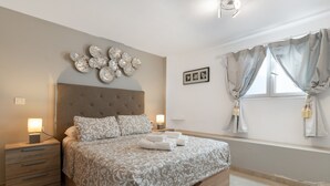 Ein Schlafzimmer mit einem großen Bett, zwei Nachttischen, einer Wanduhr, einem Fenster mit Vorhängen und einem an der Wand hängenden Bild.