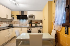 A compact kitchen with a microwave, oven, and washing machine, a dining table with chairs, and a yellow wall with a decorative item.