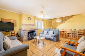 Un salon comprenant un canapé, un fauteuil, un téléviseur, une cheminée et un coin repas avec des chaises, ainsi qu’un escalier.