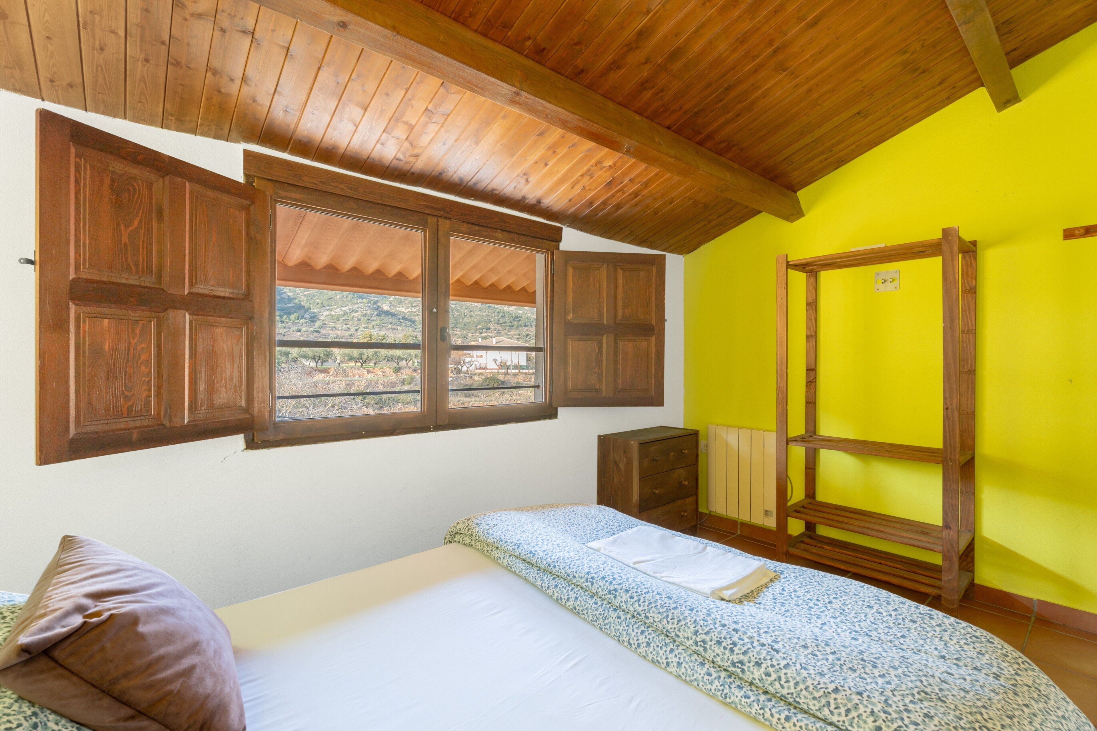 A room with wooden ceiling beams, a bed with a patterned bedspread, a window with wooden shutters, and a view of the outside.