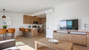 A modern kitchen with wooden cabinets, a dining area with orange chairs, a TV on a wooden stand, and a small white table.
