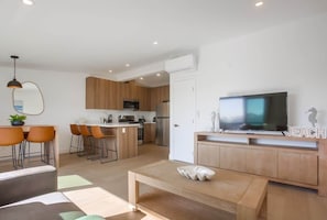 A modern kitchen with wooden cabinets, a dining area with orange chairs, a TV on a wooden stand, and a small white table.