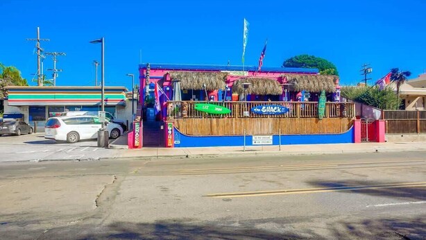 A beachfront restaurant with a thatched roof, a sign reading 'The Shack,' and a 7-Eleven store adjacent to it.
