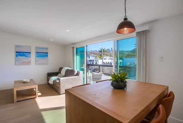 A modern living room with a wooden dining table, a sofa, and a balcony with a view of the city.