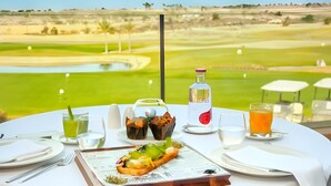 A table with a meal, drinks, and a bottle, overlooking a golf course.