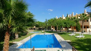 A swimming pool with a clear blue water, surrounded by palm trees and a building with multiple balconies.
