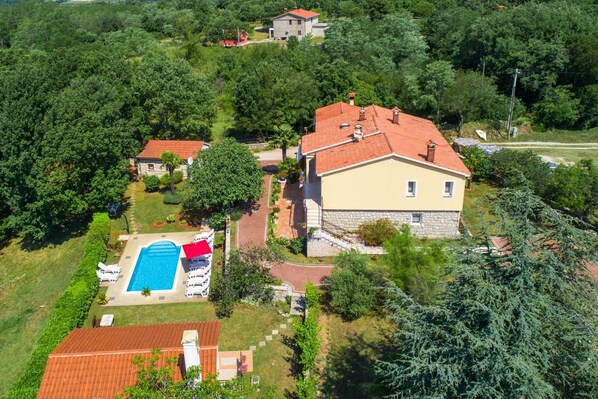 Vista aérea de una zona residencial con una casa grande que tiene piscina, rodeada de abundante vegetación y otras viviendas.