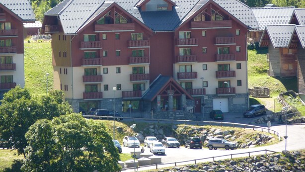 A multi-story hotel with balconies, a parking area, and a mountainous backdrop.