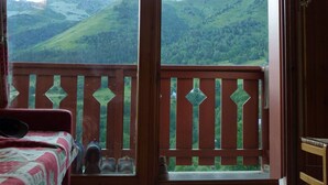 A room with a bed, a wooden balcony with a railing, and a view of green hills.