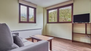 Uma sala de estar com sofá, mesa de madeira, TV em móvel e amplas janelas com vista para árvores.