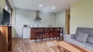 Sala de estar moderna com sofá, mesa de centro de madeira e área de cozinha com micro-ondas, pia e cadeiras.