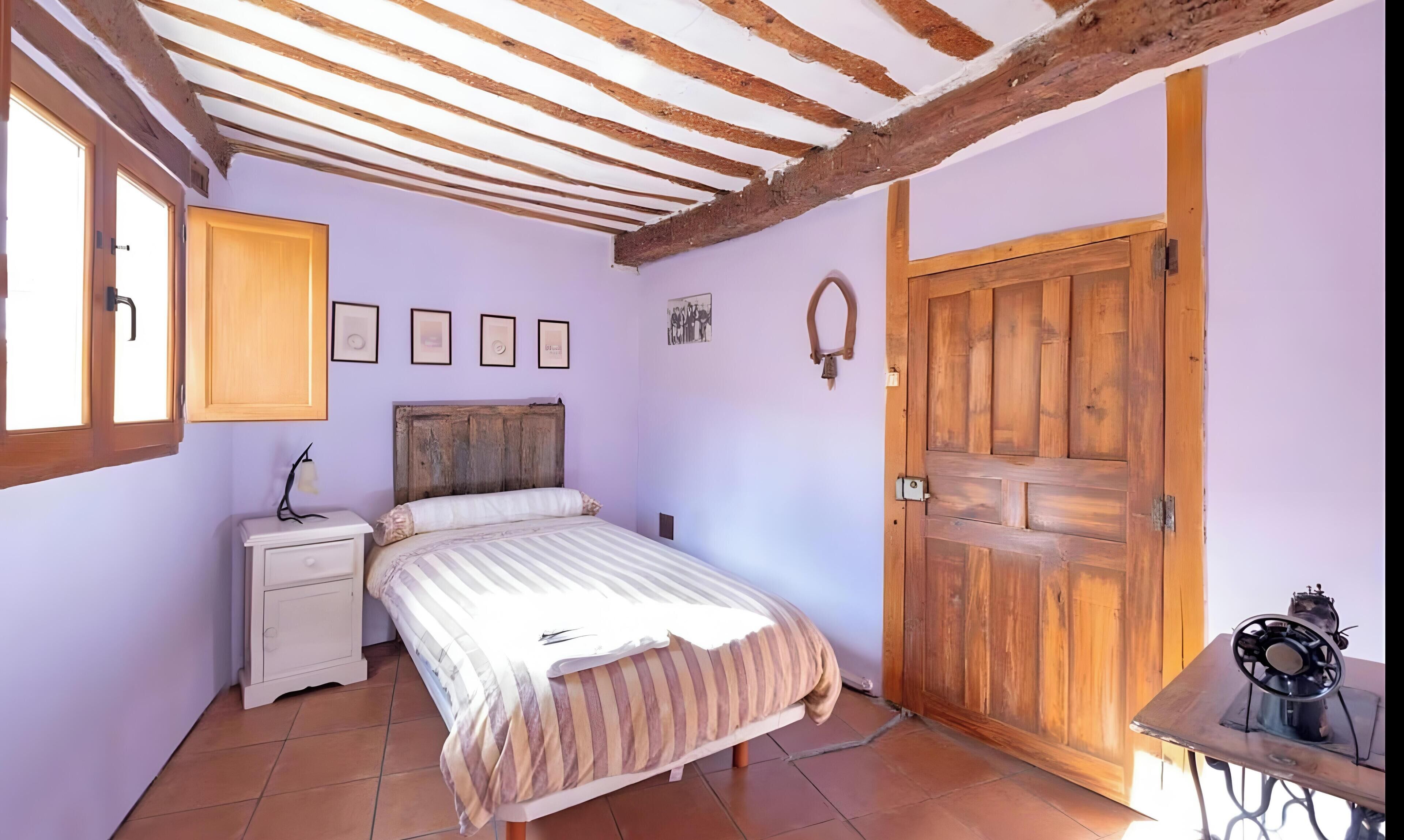 Een slaapkamer met een houten plafond, een bed met streepjesbeddengoed, een nachtkastje, een houten deur en een naaimachine op een tafel.