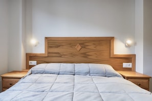 Un lit bien fait, doté d’une tête de lit matelassée, de tables de chevet en bois avec des lampes fixées au mur, et une chambre au décor simple et épuré.