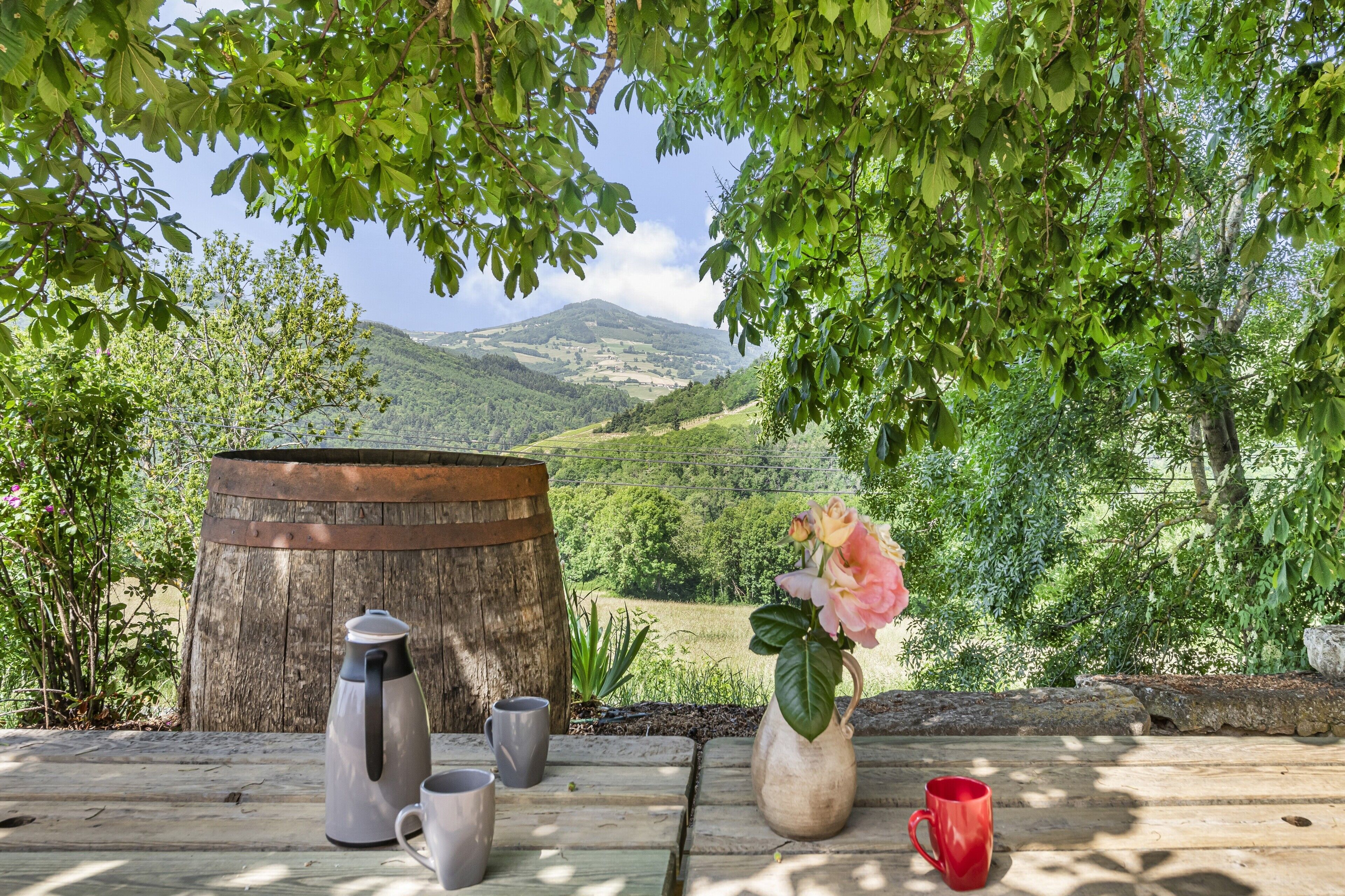 A wooden table with a coffee pot, mugs, and a vase with flowers, set against a scenic mountain backdrop.