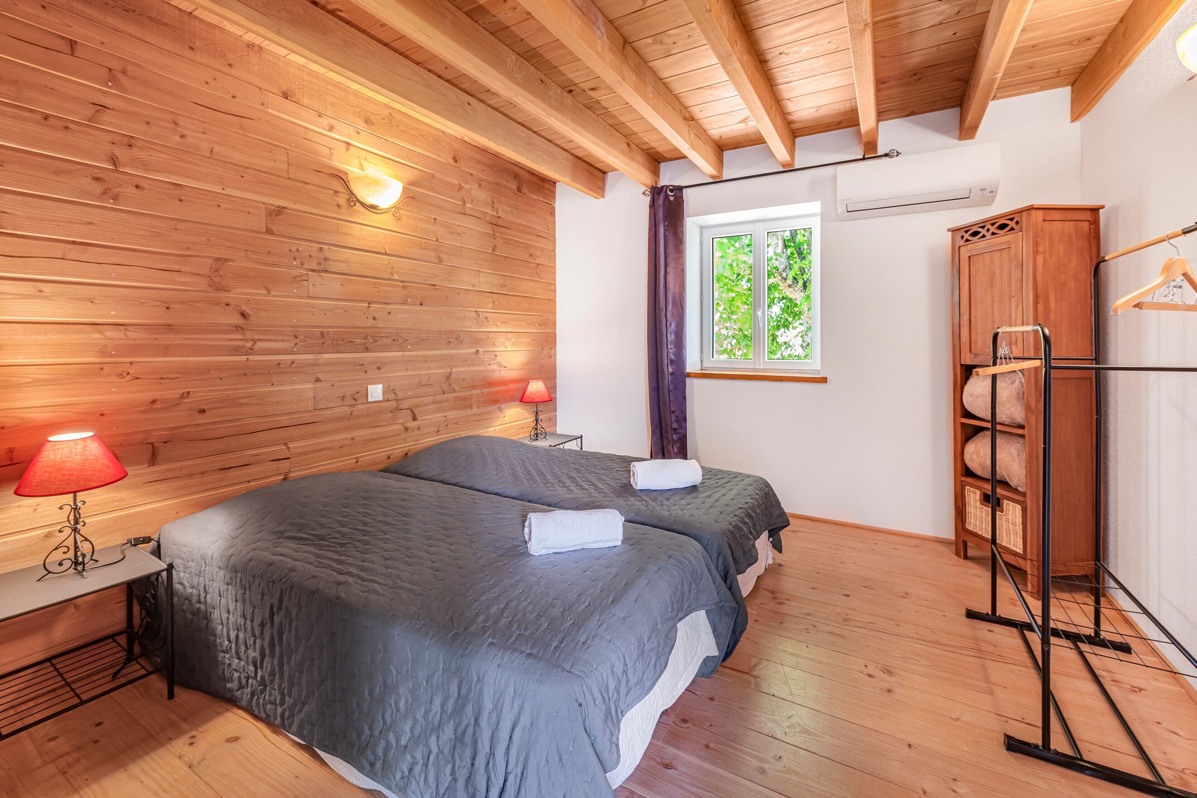 Una habitación con paredes de madera, dos camas con ropa de cama gris, un armario de madera, una ventana con vistas y una lámpara roja sobre una mesita de noche.