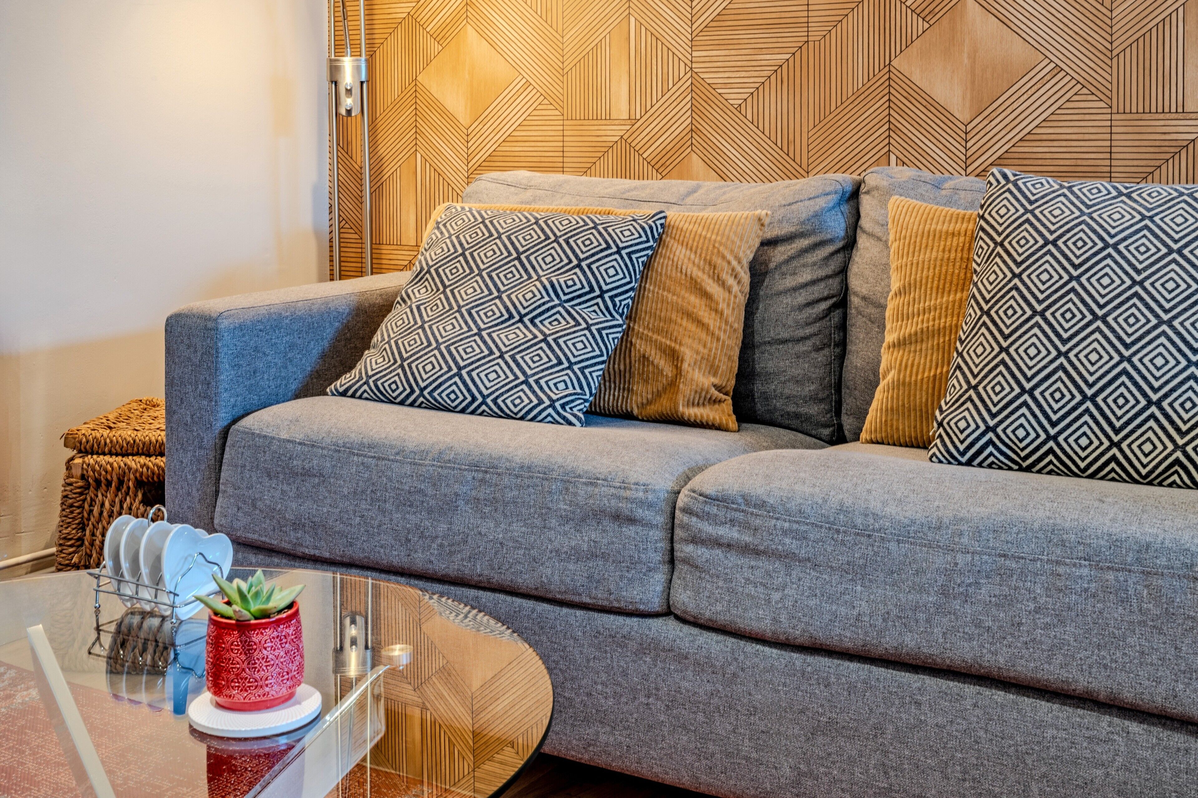 Ein modernes Wohnzimmer mit grauem Sofa, gemusterten Kissen, einem Glastisch und einer Wandverkleidung aus Holz mit geometrischem Muster.