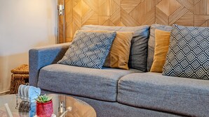 A modern living room with a grey sofa, patterned cushions, a glass coffee table, and a geometric wooden wall panel. - Studio Apartment 'Le Grand Large' with Shared Terrace, Shared Garden and Wi-Fi (Lopérec)