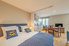Quarto com cama, almofadas, mesa de cabeceira, escrivaninha com cadeira, televisão e área de cozinha com armários e micro-ondas.