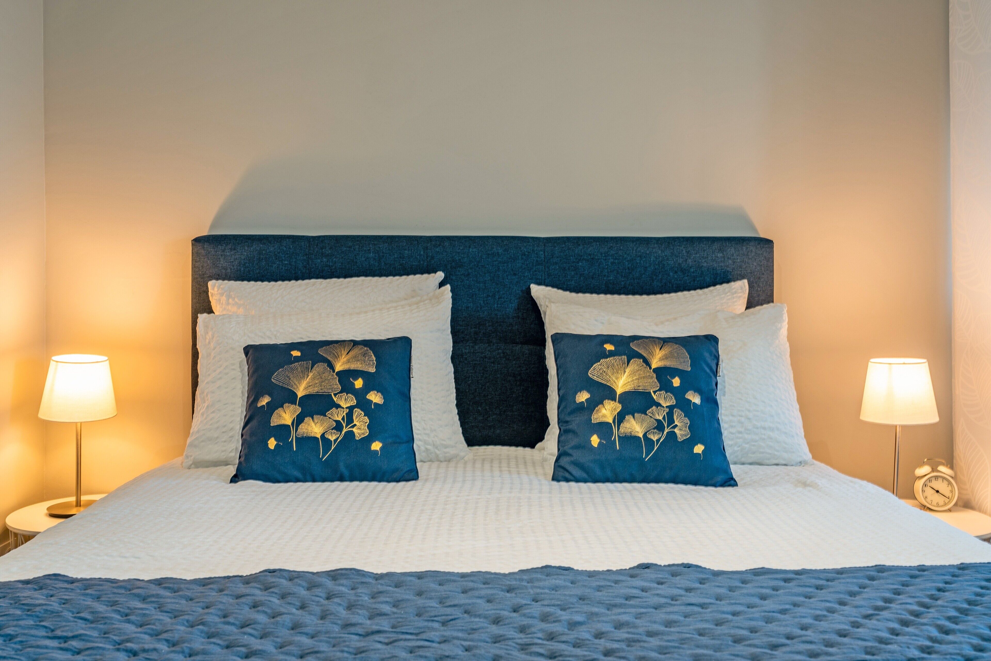 Un lit avec une tête de lit bleue, une literie blanche et deux coussins décoratifs à motifs de feuilles. On y trouve deux lampes de chevet et une horloge sur la table de chevet.