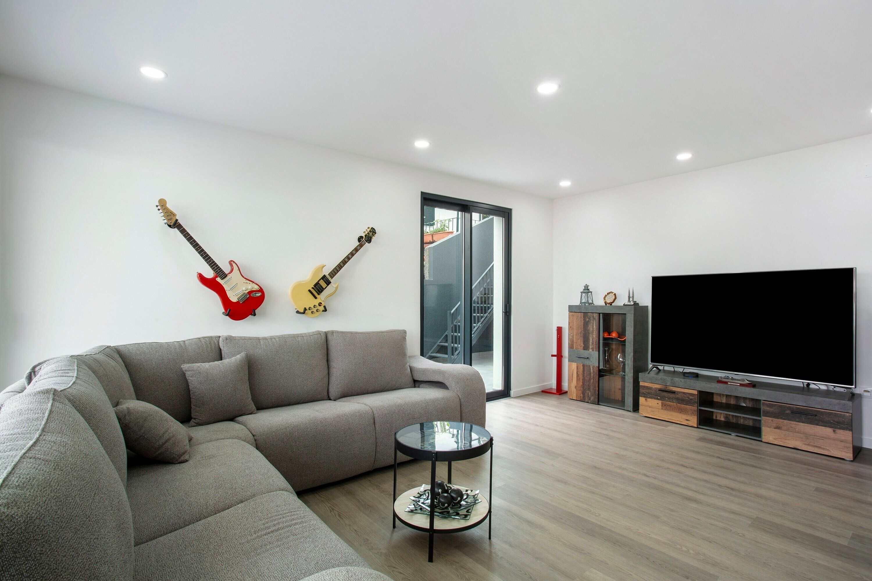 A modern living room with a grey sectional sofa, a glass coffee table, a flat-screen TV, and two guitars on the wall.