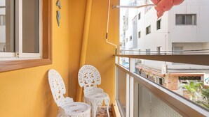 Un balcón con dos sillas blancas y una sombrilla roja, con vista a un paisaje urbano que incluye edificios y un restaurante.