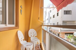 Un balcon avec deux chaises blanches et un parasol rouge, donnant sur un paysage urbain avec des immeubles et un restaurant.
