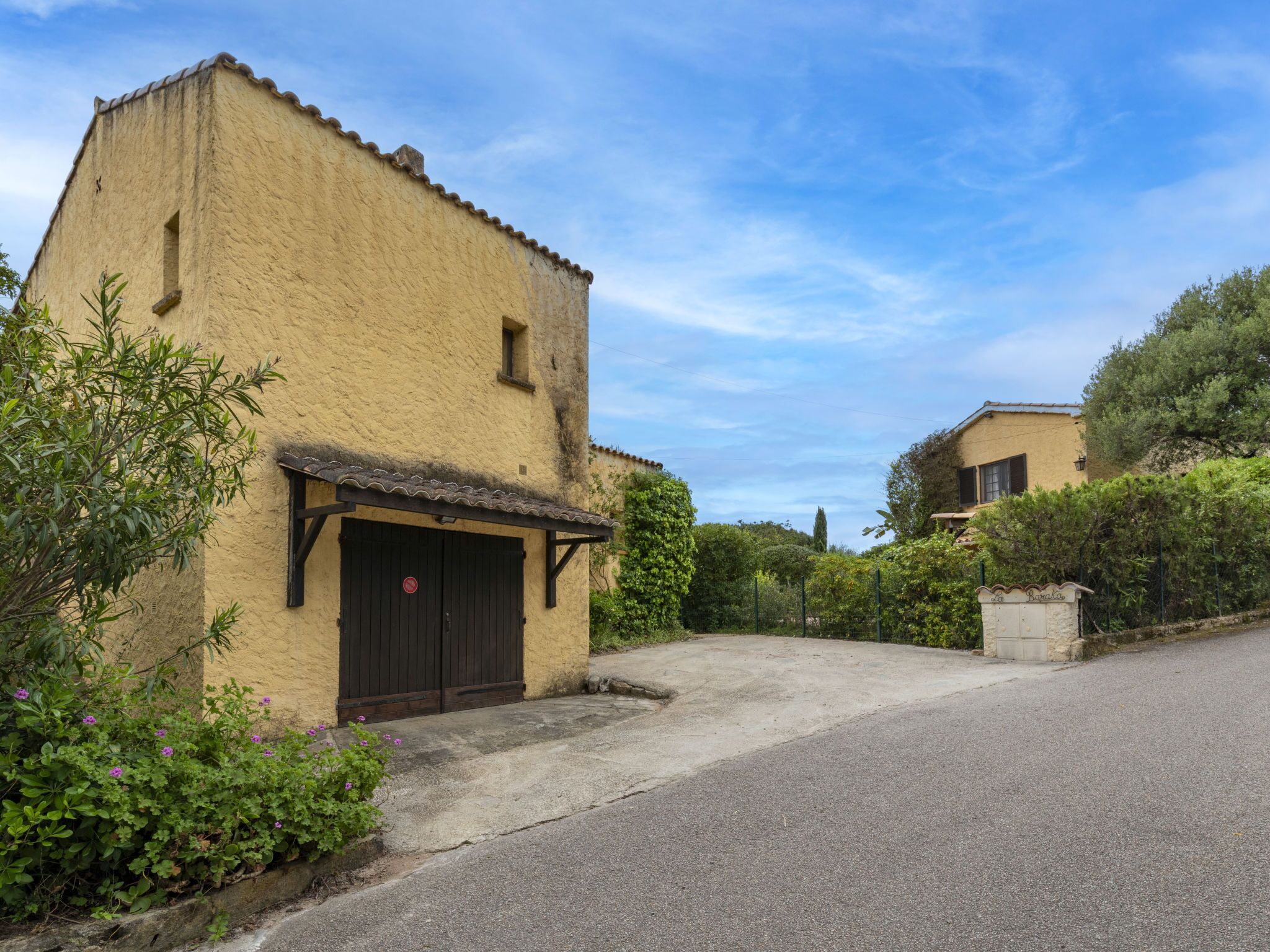 A rustic building with a flat roof and a dark door, surrounded by greenery and a paved driveway.