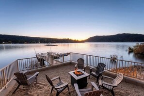 A deck with chairs and a fire pit overlooking a calm lake at sunset. - Water's edge, enjoy this lakefront property. (Big Bear Lake)