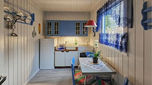 Une cuisine compacte équipée d’un réfrigérateur, d’une cuisinière et d’un évier. On y trouve une table à manger avec des chaises et une fenêtre ornée de rideaux en dentelle.