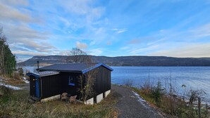 Une petite cabane avec un toit bleu, située au bord d’un lac, entourée d’arbres et sous un ciel dégagé.