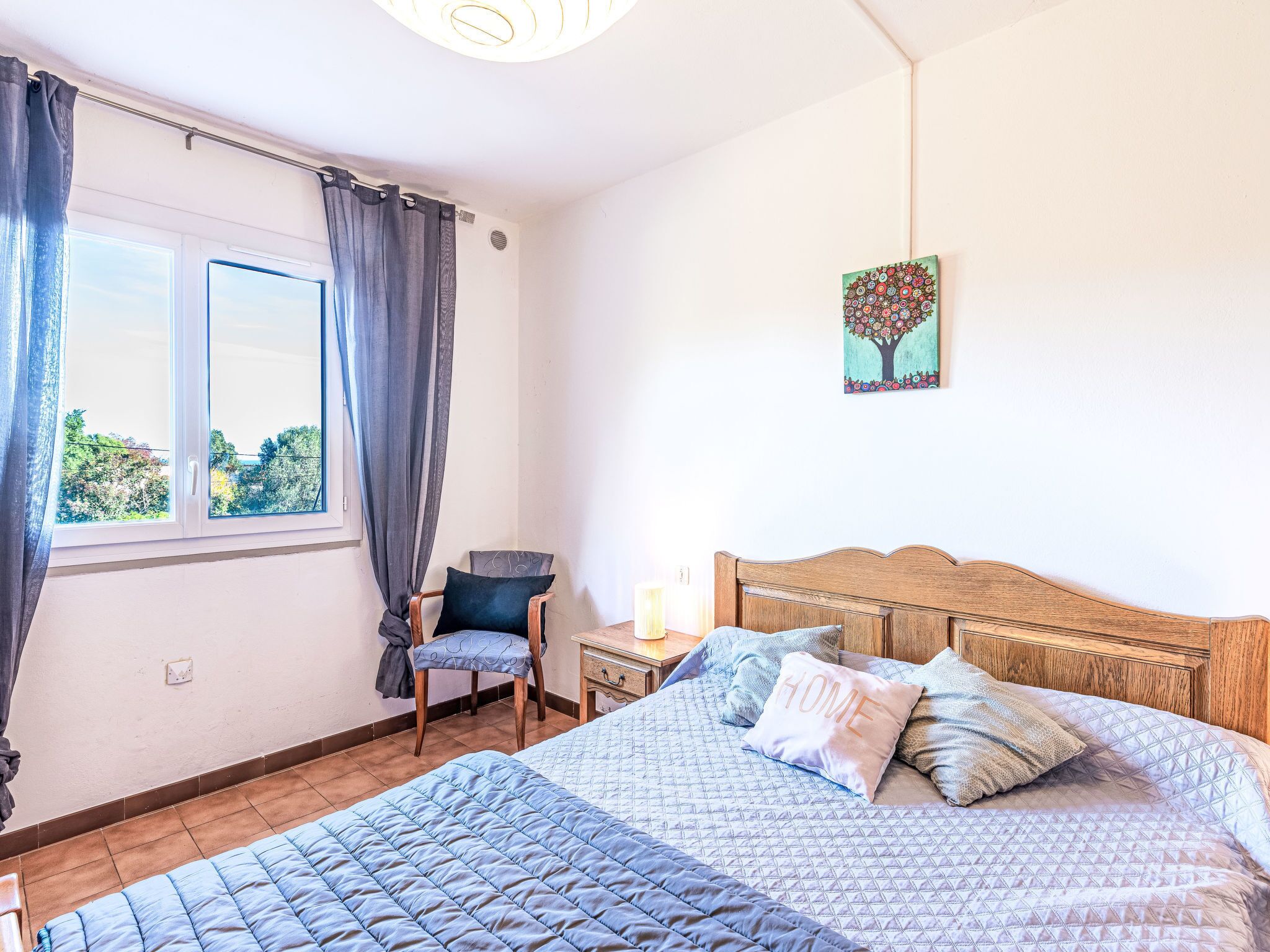 A bedroom with a bed, a chair, a nightstand, a window with curtains, and a wall painting.