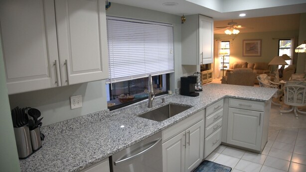 Una cocina con armarios blancos, encimera de granito, pileta de acero inoxidable y ventana con persianas.