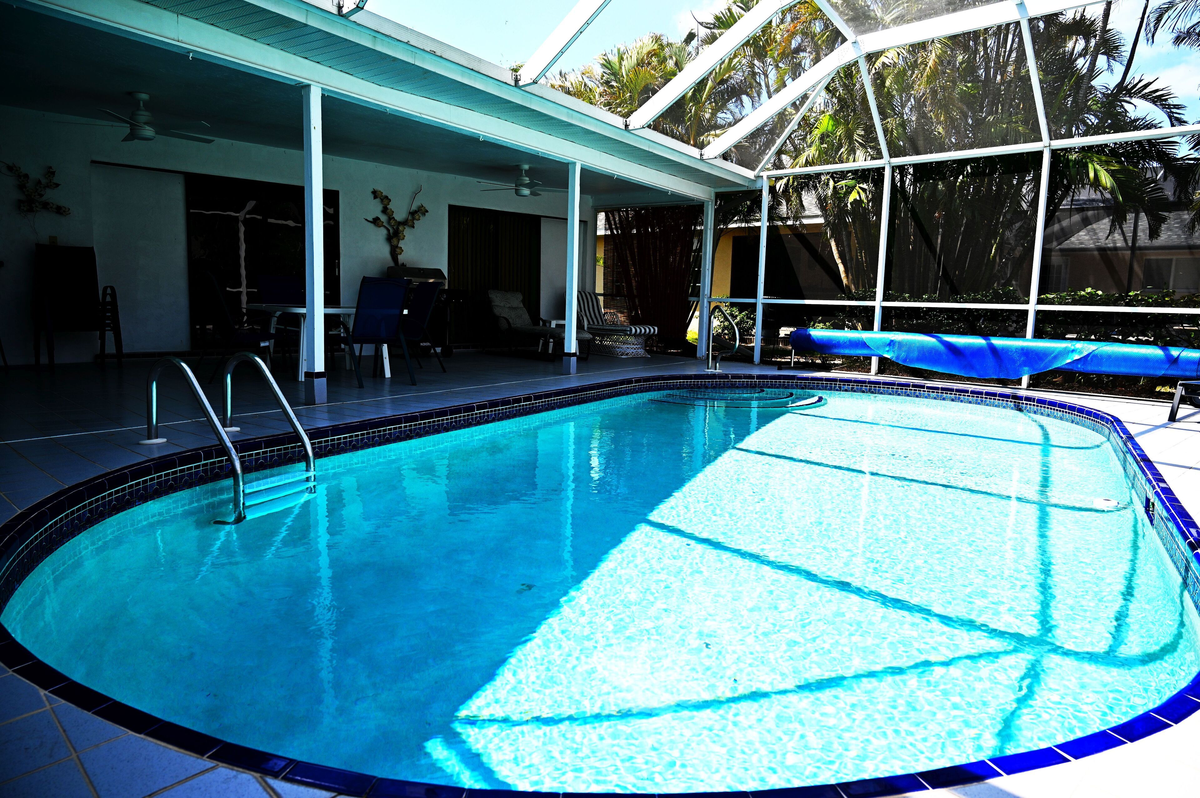 Une piscine avec un bord carrelé, un patio couvert doté de chaises et de tables, et une vue sur les palmiers à travers une paroi vitrée.