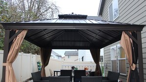 A covered patio area with wicker furniture and a table, surrounded by a white fence and a house in the background.