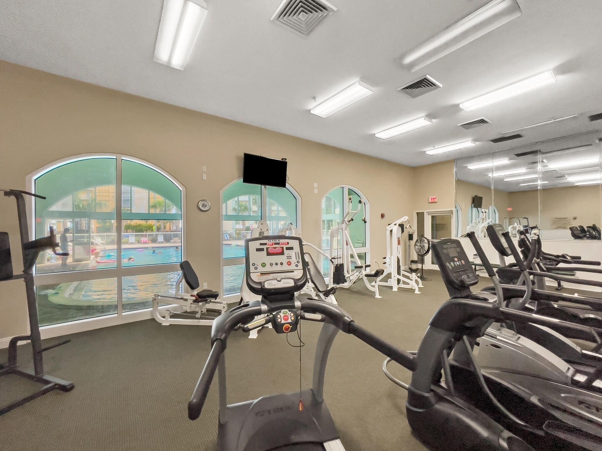 A gym with treadmills, elliptical machines, and free weights. Large windows offer views of an outdoor swimming pool.