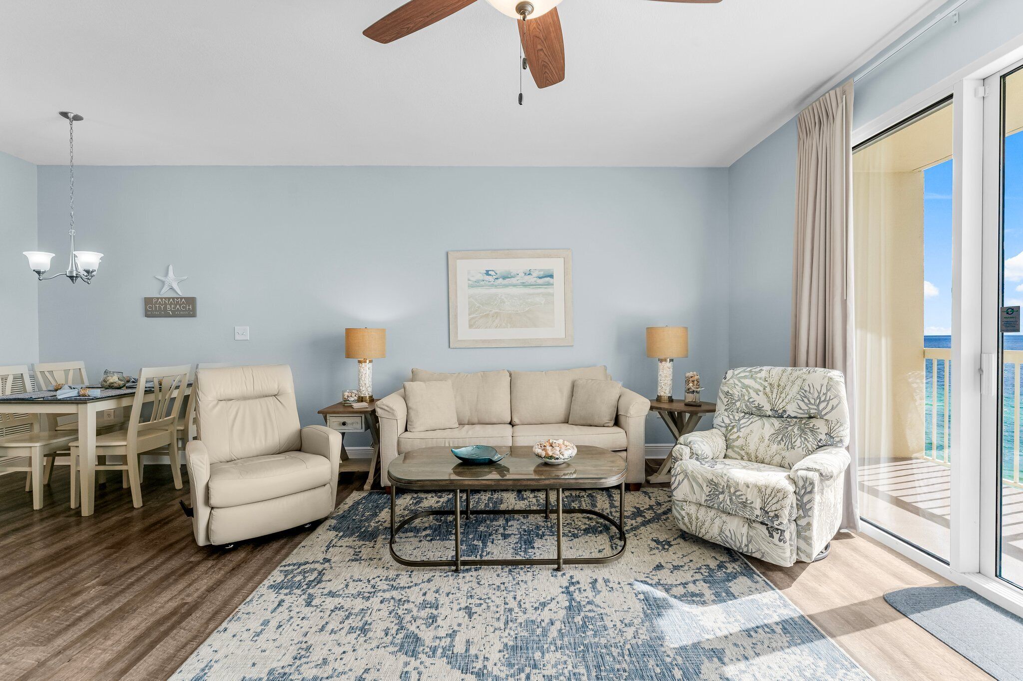 A living room with a beige sofa, a patterned armchair, a coffee table, and a dining area with a wooden table and chairs.