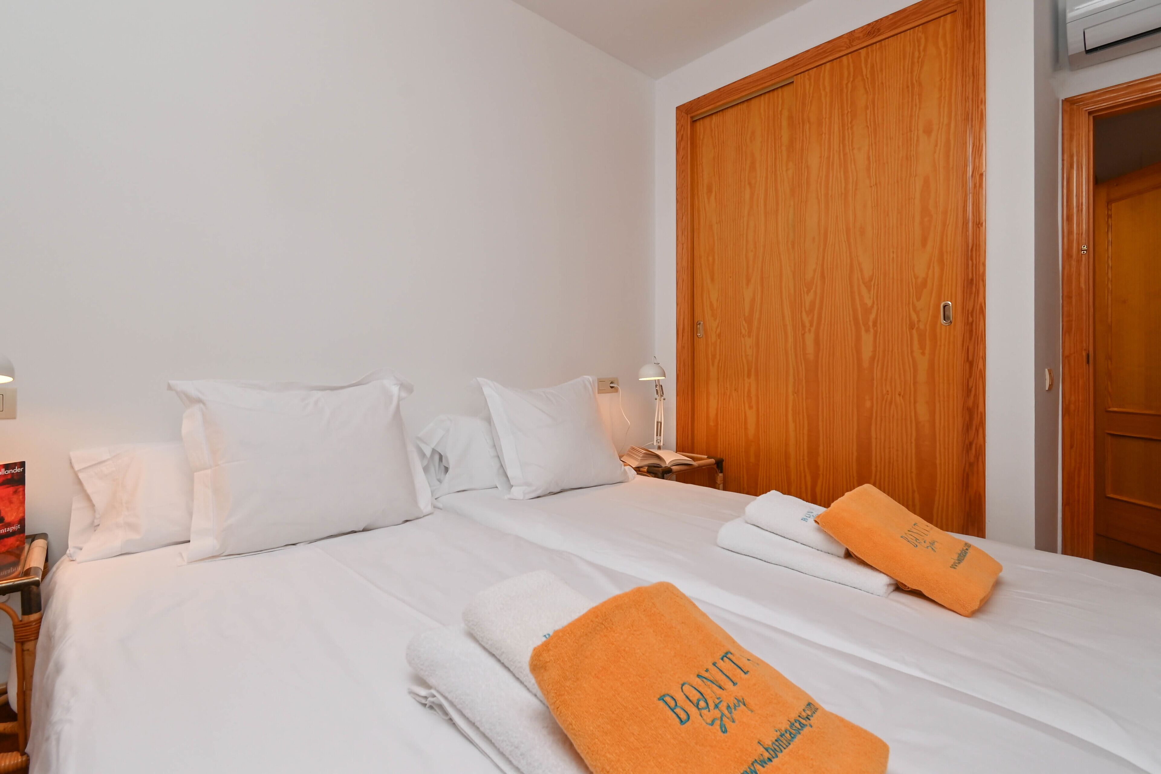 A hotel room with a large wooden wardrobe, a neatly made bed with white linens, and a pair of orange towels with text on them.