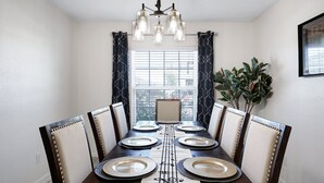 A dining room with a dark wooden table set for six, white walls, a window with curtains, and a chandelier. - Blue Note - A Birdy Vacation Rental (San Antonio)