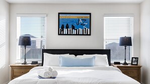 A hotel room with a large bed, two bedside tables, a framed artwork, and a view of buildings through the windows. - Blue Note - A Birdy Vacation Rental (San Antonio)