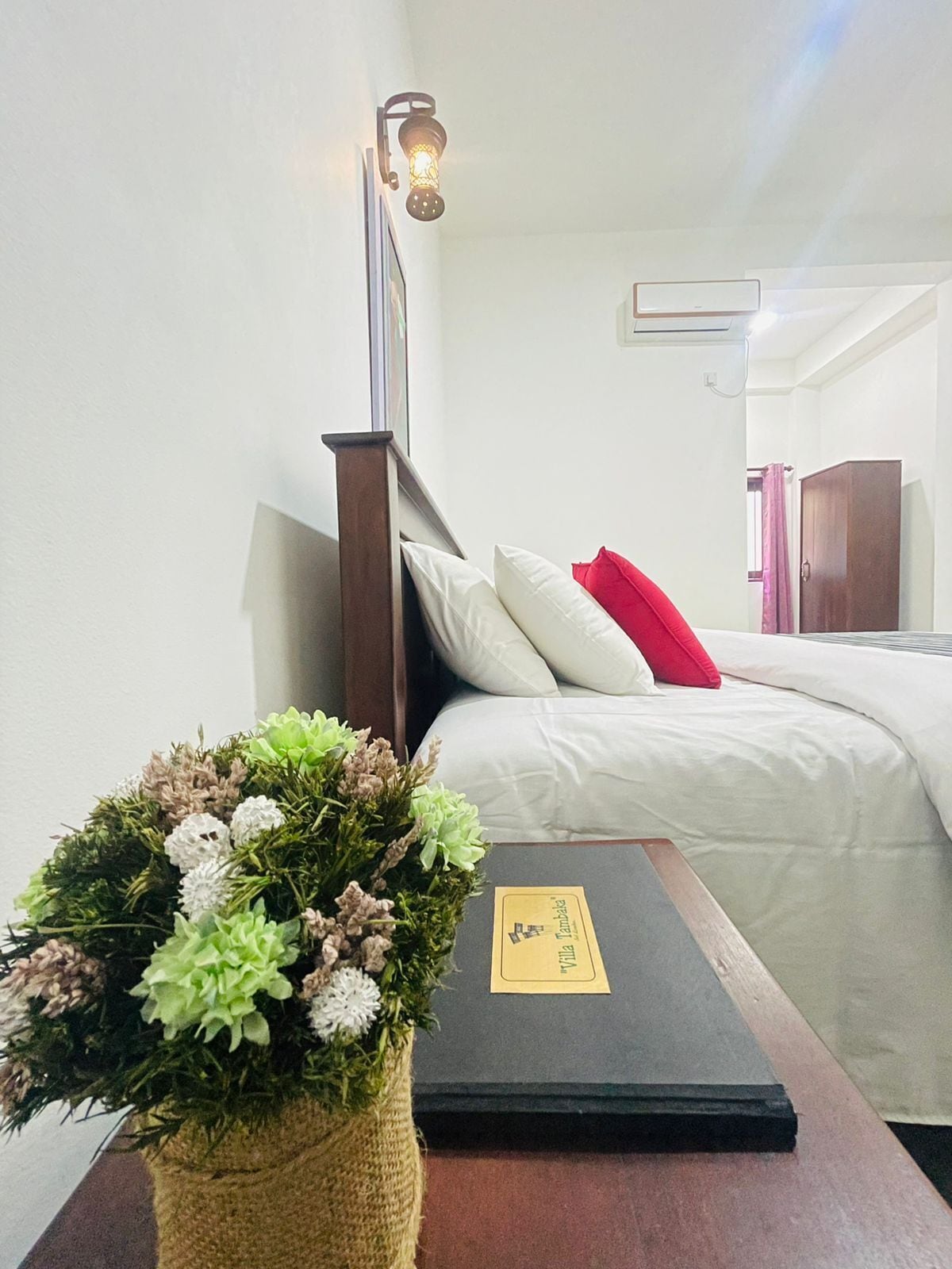 A neatly made bed with white and red pillows, a small wooden nightstand with a book and a flower arrangement, and a wall-mounted light fixture.