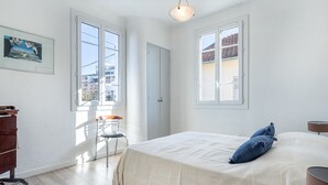 A bedroom with a large bed, two windows, a chair, a desk, and a hanging light fixture. - Phichris by Interhome (Hyères)