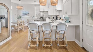 A modern kitchen with white cabinets, a central island, wicker chairs, and pendant lights. - Coast & Key Cottage by Stay on 30a (Seagrove Beach)